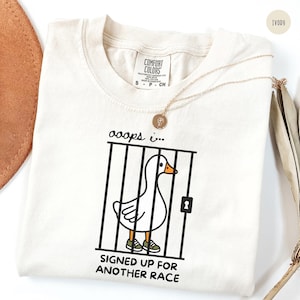 May include: An ivory t-shirt featuring a graphic of a white goose with an orange beak wearing green sneakers inside a black cage. Above the cage is the text "ooops i..." and below the cage is the text "SIGNED UP FOR ANOTHER RACE". The shirt has a Comfort Colors label.