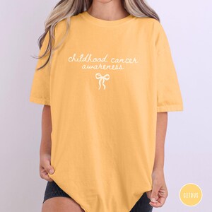 May include: A mustard yellow short-sleeve t-shirt featuring the white text "childhood cancer awareness" printed in a cursive font on the chest, accompanied by a small white line-art illustration of a ribbon bow centered below the text.