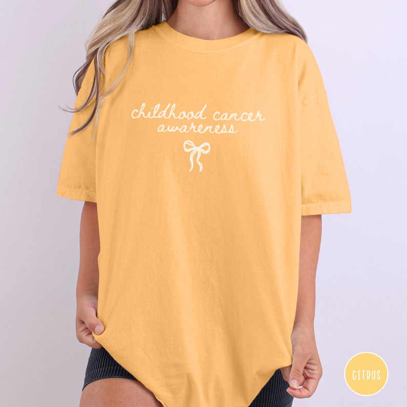 May include: A mustard yellow short-sleeve t-shirt featuring the white text "childhood cancer awareness" printed in a cursive font on the chest, accompanied by a small white line-art illustration of a ribbon bow centered below the text.