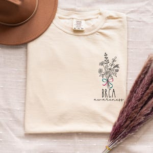 May include: Cream-colored t-shirt with a floral bouquet graphic and the text "BRCA awareness". The bouquet is tied with a pink and teal ribbon. A brown hat and dried flowers are in the background.