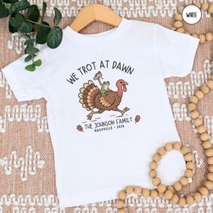 May include: A white short-sleeve t-shirt featuring a graphic of a turkey with a frog riding on its back. The text reads WE TROT AT DAWN, THE JOHNSON FAMILY, NASHVILLE, and 2026. The design includes a small sneaker held by the frog and two oak leaves.