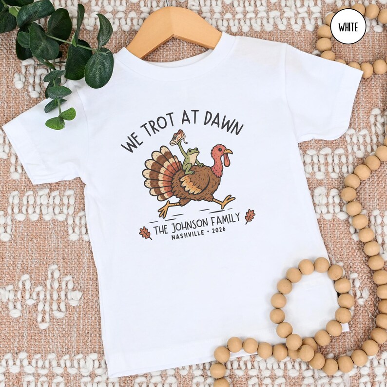 May include: A white short-sleeve t-shirt featuring a graphic of a turkey with a frog riding on its back. The text reads WE TROT AT DAWN, THE JOHNSON FAMILY, NASHVILLE, and 2026. The design includes a small sneaker held by the frog and two oak leaves.