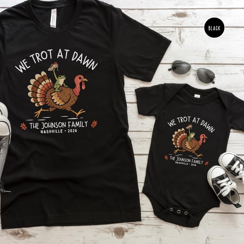 May include: A black short-sleeved t-shirt and a black short-sleeved baby bodysuit, both featuring a graphic of a turkey with a frog on its back. The text reads WE TROT AT DAWN, THE JOHNSON FAMILY, and NASHVILLE 2026, with small orange leaf accents.