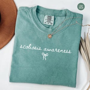 May include: A light green t-shirt with the words "scoliosis awareness" in white script and a small bow graphic. A silver necklace with a pendant rests on the shirt. The shirt is labeled "Comfort Colors".