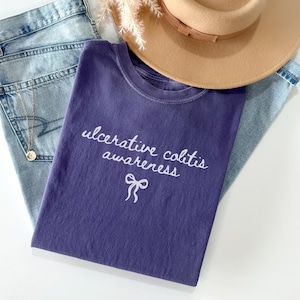 May include: A purple t-shirt with the words "ulcerative colitis awareness" and a bow graphic in white. The shirt is folded on top of light blue jeans, with a tan hat and a gold necklace.