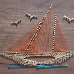 Op de afbeelding: Een zeilboot gemaakt van string art op een houten achtergrond. De zeilboot is oranje en bruin met een blauwe lijn voor het water. Er zijn drie kleine vogels gemaakt van string art op de achtergrond.