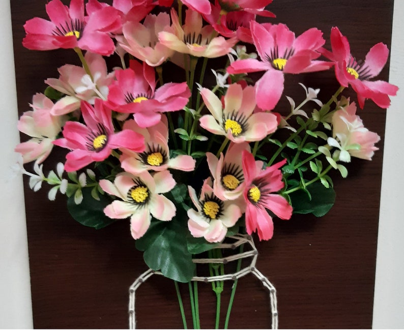 May include: A bouquet of pink and white flowers with green stems arranged in a wire jar. The flowers are in a wooden frame.
