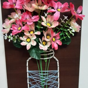 May include: A string art piece depicting a mason jar filled with pink and white flowers. The jar is made of blue and white string wrapped around nails hammered into a wooden board.