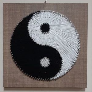 May include: A black and white string art piece depicting the yin yang symbol on a wood background.