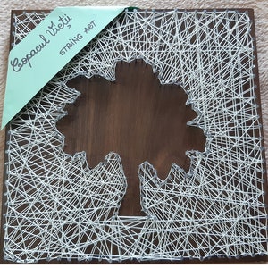 Peut inclure: Une œuvre d'art en fil de bois carrée représentant un arbre. Du fil blanc est tissé autour de clous pour créer l'arbre et l'arrière-plan. L'arbre est placé sur un fond marron foncé. Les mots "Copacul Vieții" et "STRING ART" sont sur une étiquette verte.