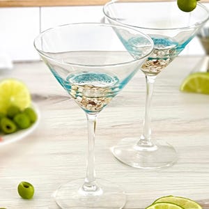 May include: Two clear glass martini glasses with blue and gold accents. The glasses are filled with a clear liquid and garnished with green olives. The glasses are sitting on a white wooden surface with lime slices and green olives.