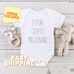 May include: White baby onesie with the text "Future Crypto Millionaire" printed in black. The onesie is on a wooden surface with a stuffed animal and a wooden teether.