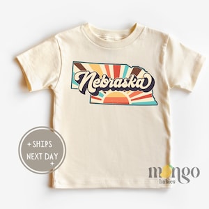 May include: A cream colored short-sleeved t-shirt with a retro-style graphic of the state of Nebraska with a sunburst design and the word "Nebraska" in a vintage font.