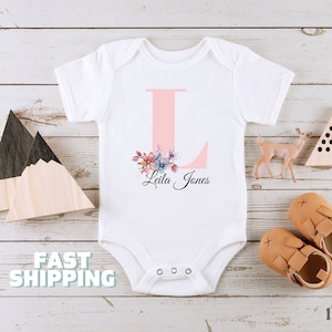 May include: White baby onesie with a pink letter L and floral design. The onesie is personalized with the name "Leila Jones".