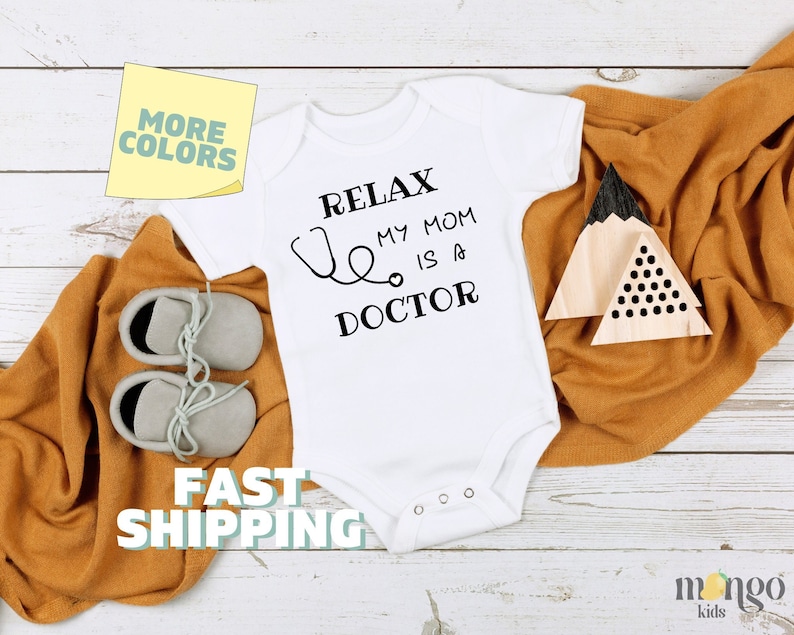 Op de afbeelding: Witte baby romper met zwarte tekst die "Relax My Mom Is A Doctor" zegt en een stethoscoop afbeelding. De romper ligt op een wit houten oppervlak met een bruine deken en een houten speelgoedberg.