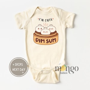 May include: A cream-colored baby onesie with short sleeves features a cartoon illustration of three steamed buns with happy faces in a bamboo steamer. The text "I'm Cute" and "Dim Sum" are printed on the onesie.