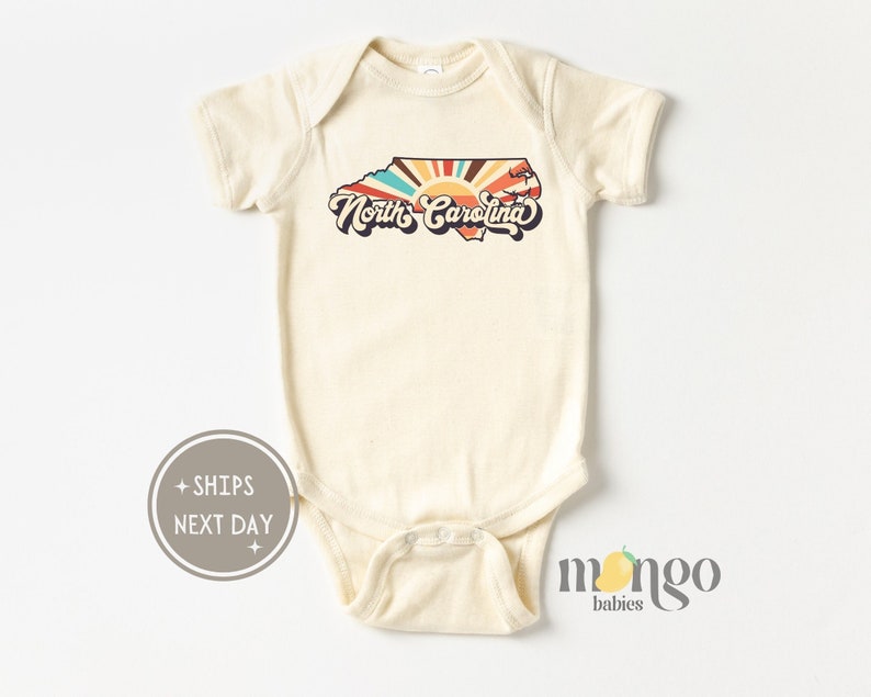 Puede incluir: Un body de beb&eacute; de color crema con un gr&aacute;fico retro del estado de Carolina del Norte con un dise&ntilde;o de rayos de sol y el texto "North Carolina".