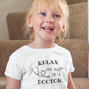 Op de afbeelding: Een wit T-shirt met een zwarte afbeelding waarop staat "Relax My Mom Is A Doctor" met een stethoscoop. Een jong meisje met blond haar draagt het shirt en glimlacht.