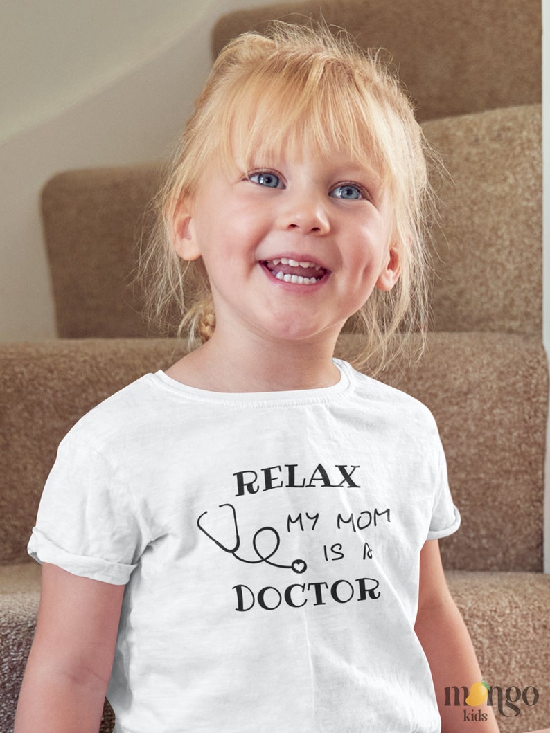 Op de afbeelding: Een wit T-shirt met een zwarte afbeelding waarop staat "Relax My Mom Is A Doctor" met een stethoscoop. Een jong meisje met blond haar draagt het shirt en glimlacht.