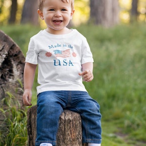 May include: A young child wearing a white t-shirt with a red, white, and blue American flag graphic and the text "Made in the USA". The child is sitting on a tree stump in a grassy area.