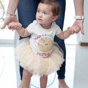 K&ouml;nnte beinhalten: Ein kleines M&auml;dchen tr&auml;gt einen wei&szlig;en Strampler mit dem Text "Mama's Little Pupus" und einem braunen Keks-Motiv. Sie tr&auml;gt einen beigefarbenen T&uuml;llrock mit goldenen Glitzerpunkten und h&auml;lt die Hand eines Erwachsenen.