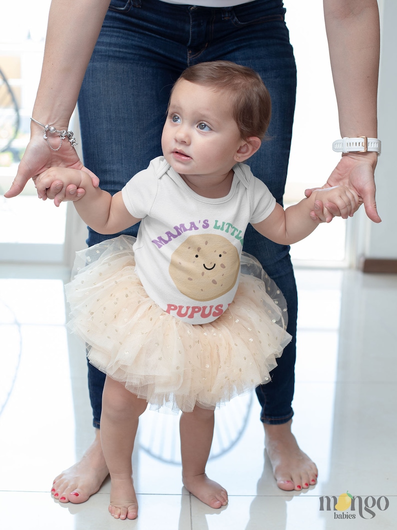 K&ouml;nnte beinhalten: Ein kleines M&auml;dchen tr&auml;gt einen wei&szlig;en Strampler mit dem Text "Mama's Little Pupus" und einem braunen Keks-Motiv. Sie tr&auml;gt einen beigefarbenen T&uuml;llrock mit goldenen Glitzerpunkten und h&auml;lt die Hand eines Erwachsenen.