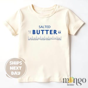 May include: Cream-colored t-shirt with the words "SALTED BUTTER" in blue, along with a measurement guide. The shirt also includes the text "NET WT 4 OZ (113G)" and "GRADE AA".
