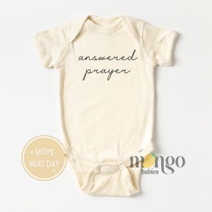 May include: Cream-colored baby onesie with the words "answered prayer" in a delicate, cursive font. The onesie has short sleeves and a snap closure. The image also includes a logo and a circular sticker.