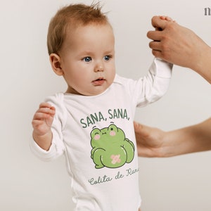 K&ouml;nnte beinhalten: Ein wei&szlig;er Langarm-Strampler mit einem gr&uuml;nen Frosch-Motiv und dem Text "Sana, Sana, Colita de Rana".