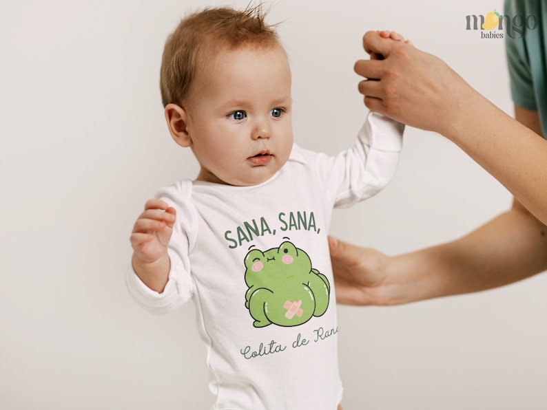 K&ouml;nnte beinhalten: Ein wei&szlig;er Langarm-Strampler mit einem gr&uuml;nen Frosch-Motiv und dem Text "Sana, Sana, Colita de Rana".