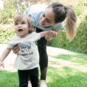 May include: A young girl wearing a white t-shirt with the text "I have the best grandma ever" and a small elephant graphic. She is holding hands with a woman and smiling.