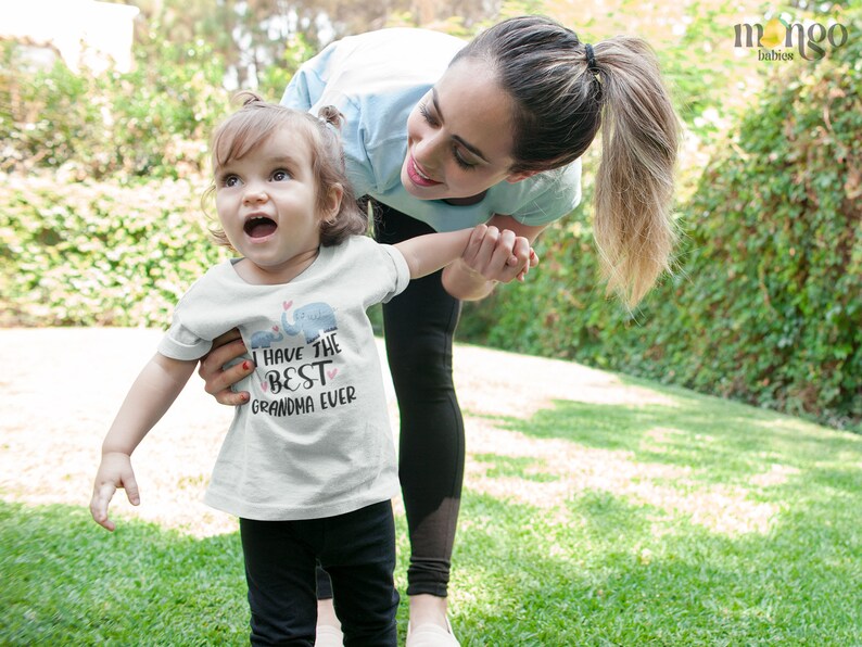 May include: A young girl wearing a white t-shirt with the text "I have the best grandma ever" and a small elephant graphic. She is holding hands with a woman and smiling.