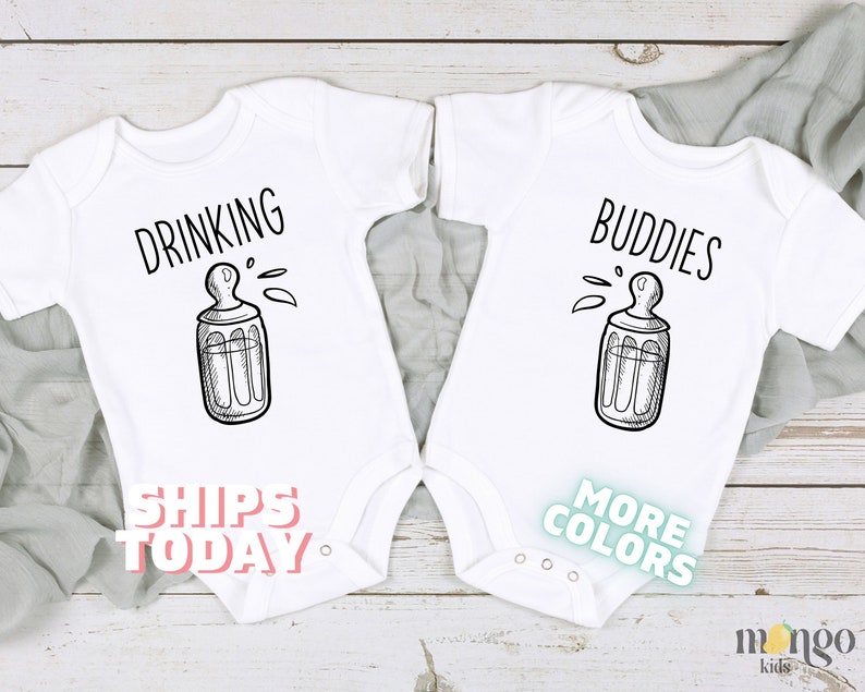 May include: Two white baby onesies with black text and a drawing of a baby bottle. The onesie on the left says "Drinking" and the onesie on the right says "Buddies".