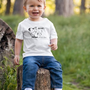 Może przedstawiać: Małe dziecko w białej koszulce z czarno-białym rysunkiem osoby śpiewającej z tekstem "MAMA! OO-OO-OO-OH!" Dziecko ma na sobie niebieskie dżinsy i siedzi na pniu drzewa w trawiastym miejscu.