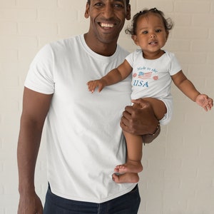 May include: A father and daughter wearing matching white t-shirts. The daughter's shirt has a red, white, and blue American flag design with the text "Made in the USA".