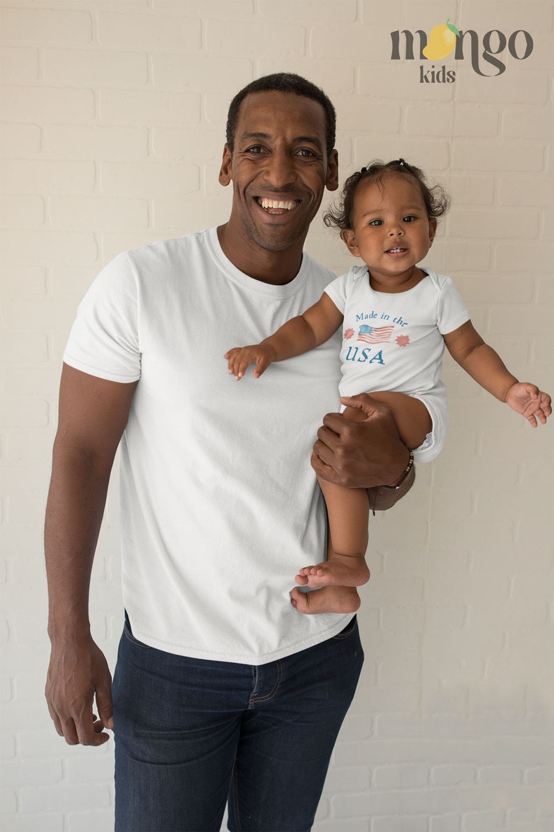 May include: A father and daughter wearing matching white t-shirts. The daughter's shirt has a red, white, and blue American flag design with the text "Made in the USA".