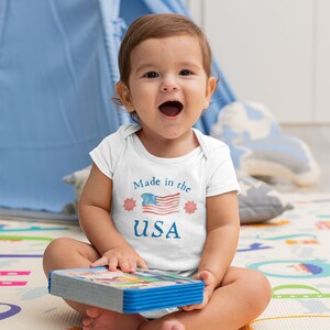 May include: A white baby onesie with a blue and red watercolor American flag and the text "Made in the USA". A baby is sitting on a colorful play mat holding a book.