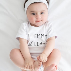Peut inclure: Un body blanc pour b&eacute;b&eacute; avec du texte noir qui dit "Emma" et "Leah" avec une coche &agrave; c&ocirc;t&eacute; de Leah. Le body est sur un fond blanc.