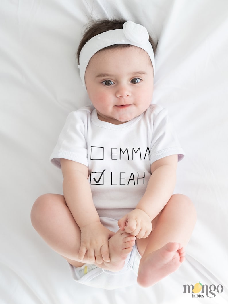 Peut inclure: Un body blanc pour b&eacute;b&eacute; avec du texte noir qui dit "Emma" et "Leah" avec une coche &agrave; c&ocirc;t&eacute; de Leah. Le body est sur un fond blanc.