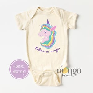 May include: A cream-colored baby onesie with a colorful unicorn illustration and the text "believe in magic".