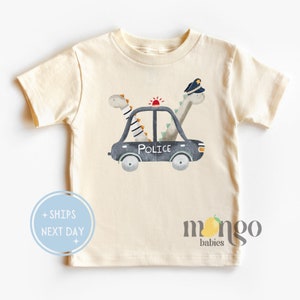 May include: A light beige short-sleeved t-shirt with a graphic of two dinosaurs in a police car. The car has a red light on top and the word "POLICE" written on the side. The text "mango babies" is printed on the bottom right corner of the shirt.