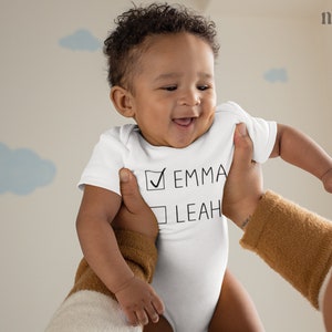 Peut inclure: Un body blanc pour b&eacute;b&eacute; avec une liste de contr&ocirc;le sur le devant. La liste de contr&ocirc;le comporte deux options&nbsp;: Emma et Leah. La case &agrave; c&ocirc;t&eacute; d&rsquo;Emma est coch&eacute;e.