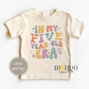 May include: A light beige t-shirt with a colorful graphic that says "In My Five Year Old Era" with flowers and a butterfly. The text is in a retro font style.