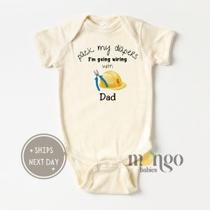 May include: Cream-colored baby onesie with the text "pack my diapers I'm going wiring with Dad." The onesie features a graphic of a yellow hard hat, blue pliers, and a red lightning bolt. The onesie is designed for infants.