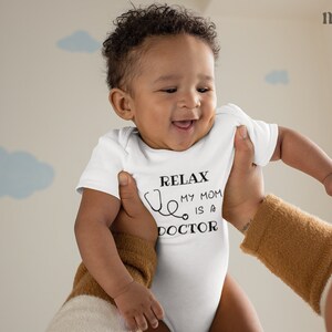 Op de afbeelding: Een witte baby romper met zwarte tekst die "Relax, My Mom is a Doctor" luidt met een stethoscoop afbeelding. Een baby wordt omhoog gehouden door een volwassene.