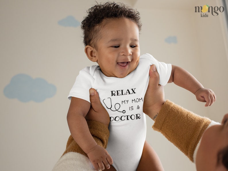 Op de afbeelding: Een witte baby romper met zwarte tekst die "Relax, My Mom is a Doctor" luidt met een stethoscoop afbeelding. Een baby wordt omhoog gehouden door een volwassene.