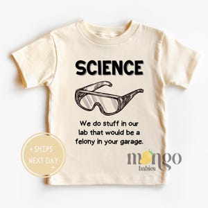 May include: A beige t-shirt with the text "SCIENCE" in black letters. The shirt also features a drawing of safety goggles and the text "We do stuff in our lab that would be a felony in your garage."