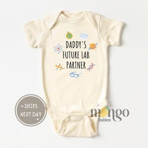 May include: A cream-colored baby onesie with the text "Daddy's Future Lab Partner" and science-themed illustrations, including a beaker, atom, DNA strand, and planets.