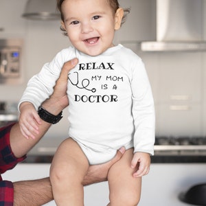 Op de afbeelding: Een witte baby romper met lange mouwen met zwarte tekst die "Relax, My Mom is a Doctor" luidt en een stethoscoop afbeelding.