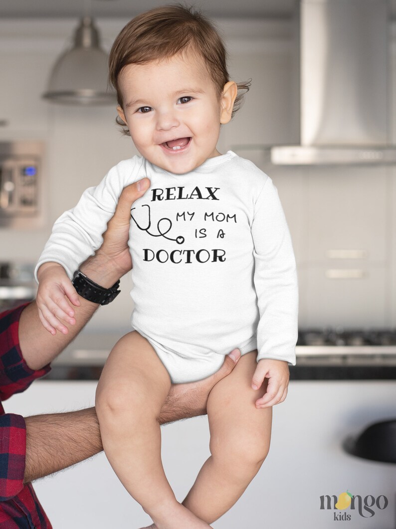Op de afbeelding: Een witte baby romper met lange mouwen met zwarte tekst die "Relax, My Mom is a Doctor" luidt en een stethoscoop afbeelding.